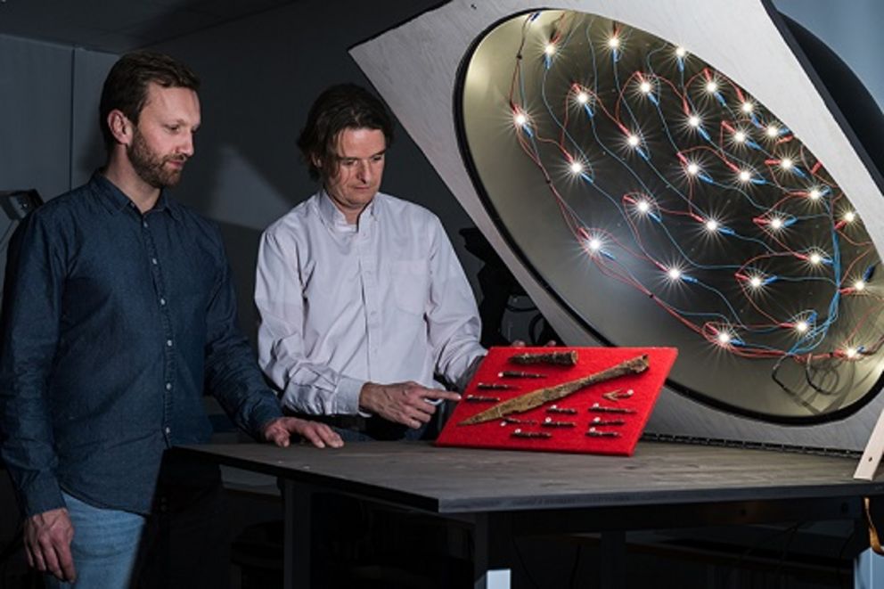 Prof. Dr. Malte Rehbein und Sebastian Gassner stehen neben der RTI-Apparatur und schauen auf mittelalterliche Waffenteile, die sie digitalisieren möchten.